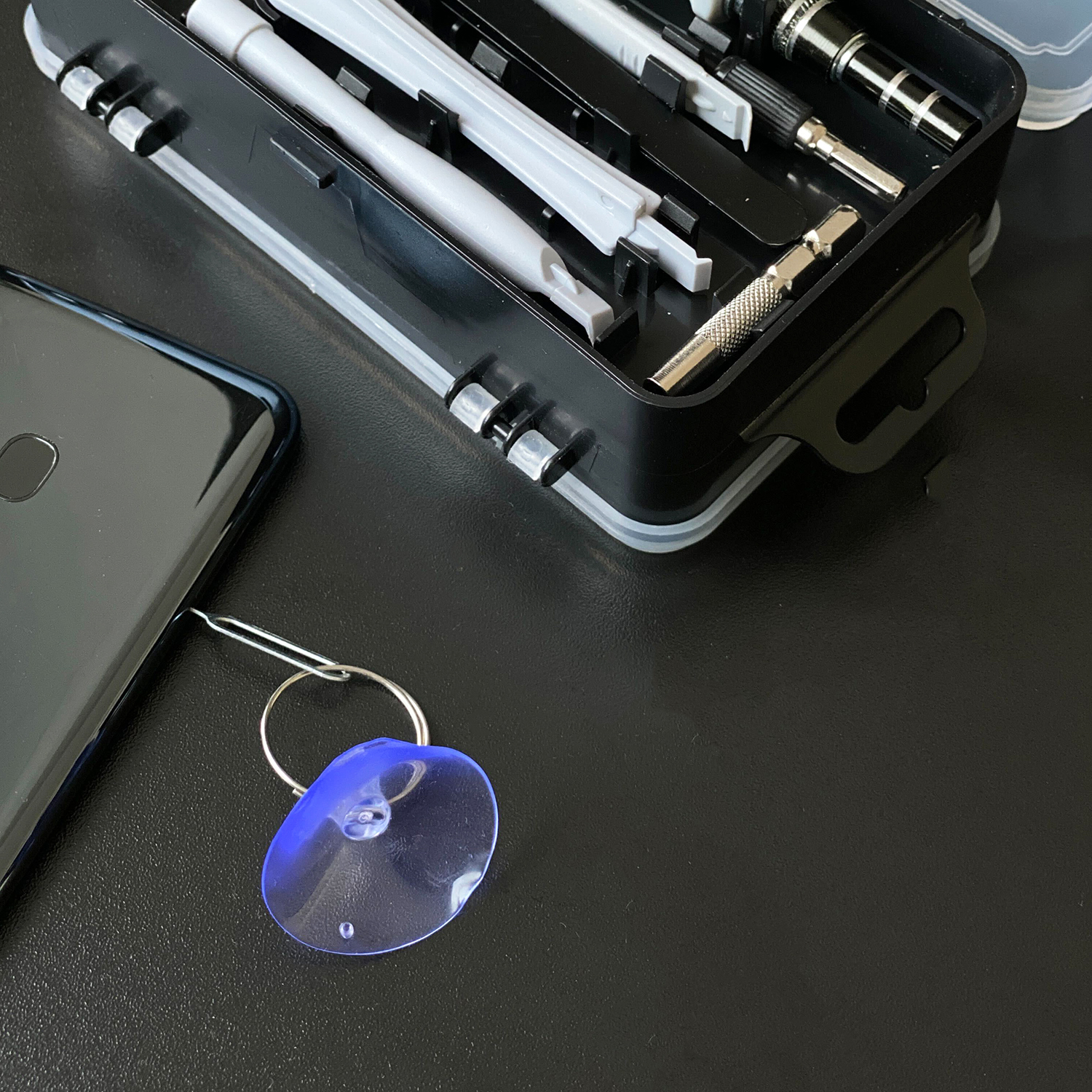 Ein Saugnapf und Werkzeuge liegen auf einer schwarzen Oberfläche. Ein schwarzes Telefon in der Ecke.