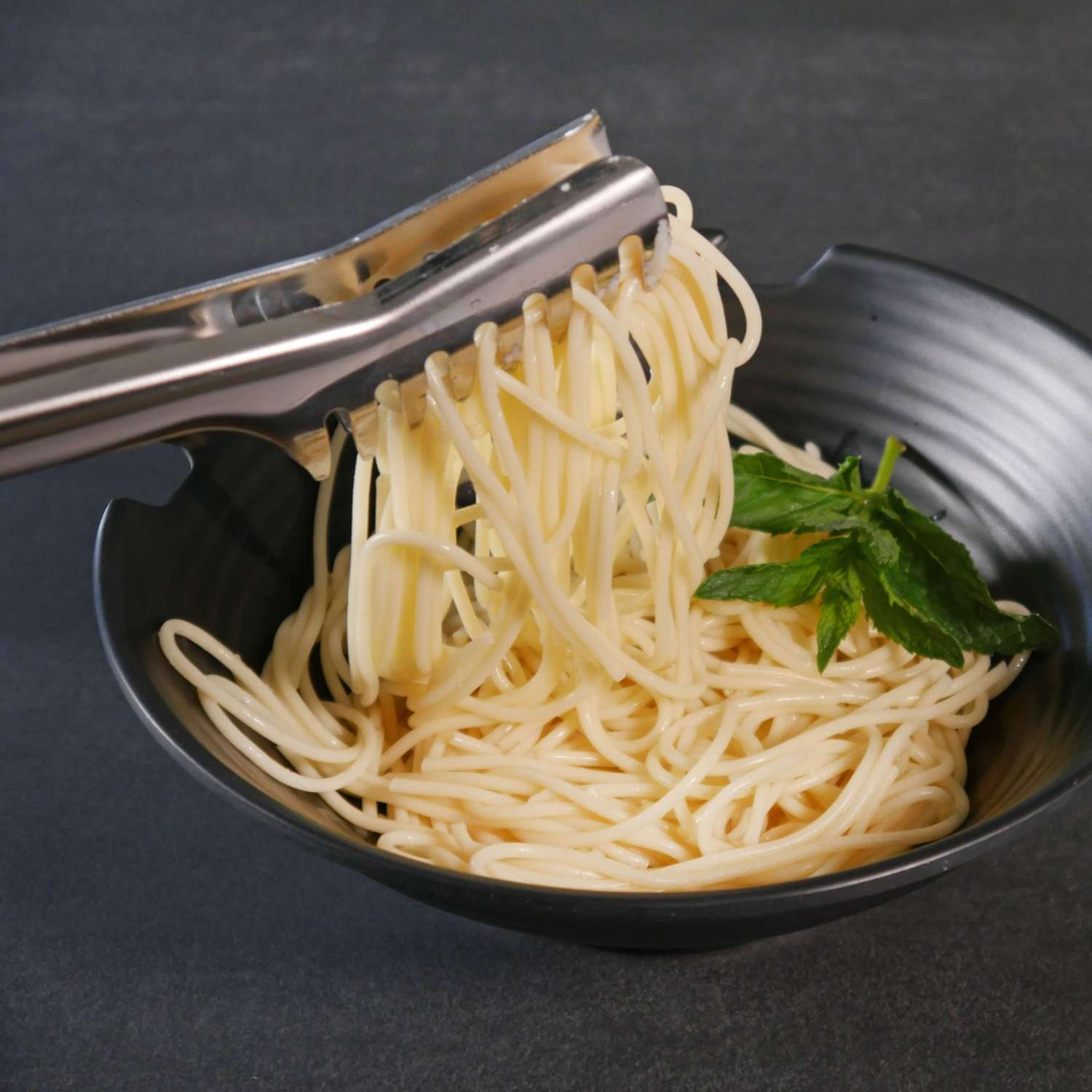 Spaghetti-Nudeln werden mit Metallzangen aus einer schwarzen Schüssel geholt, mit grünem Kraut daneben.