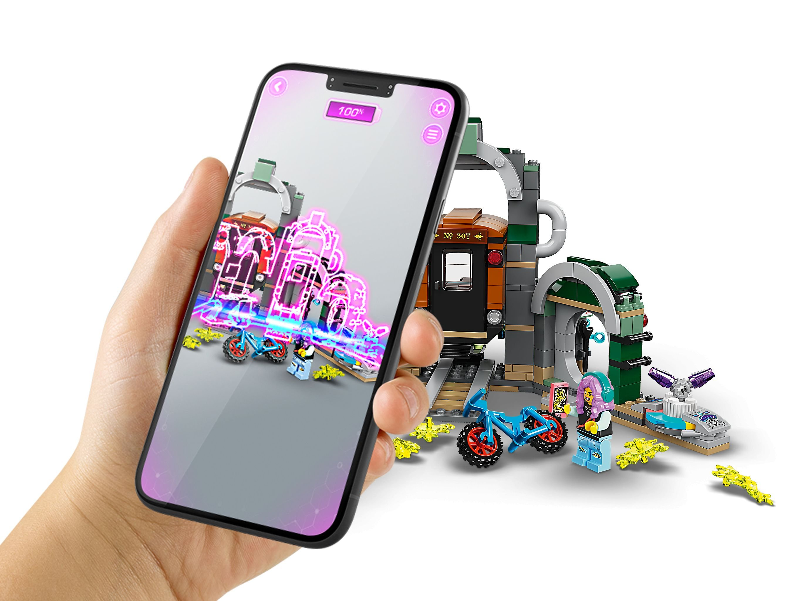 Ręka trzyma telefon pokazujący scenę z Lego, stację kolejową i figurki. Białe tło.