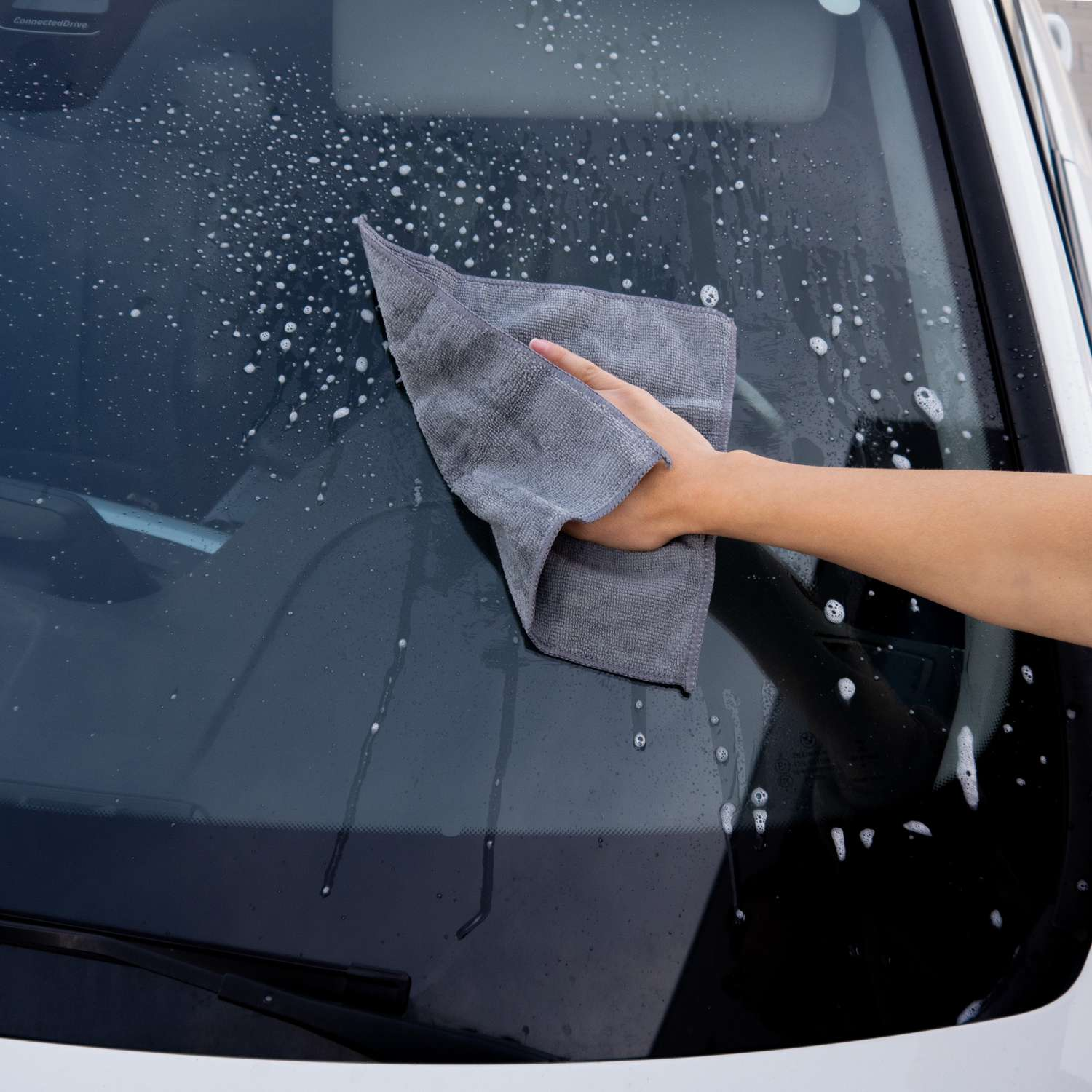 Eine Hand wischt eine Seifenblasen-Autoscheibe mit einem grauen Tuch ab.