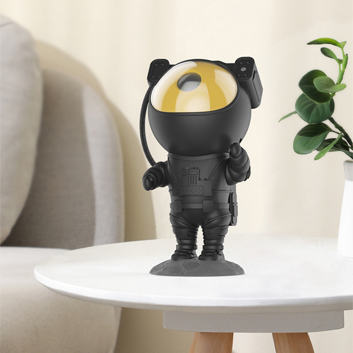 Eine schwarz astronautenförmige Lampe auf einem weißen Tisch, mit einer weißen Vase und einem grauen Sofa im Hintergrund.