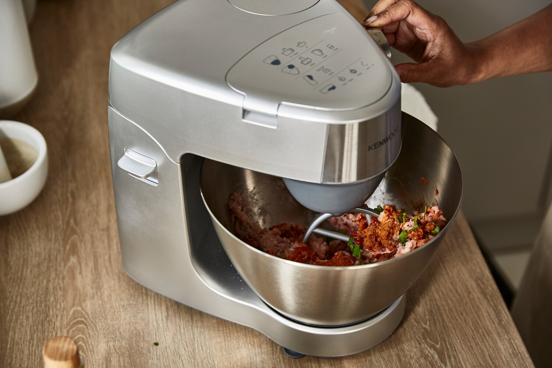 Ein silberner Kenwood-Küchenmixer, der Zutaten in einer Schüssel vermischt. Eine Hand stellt die Einstellungen ein.