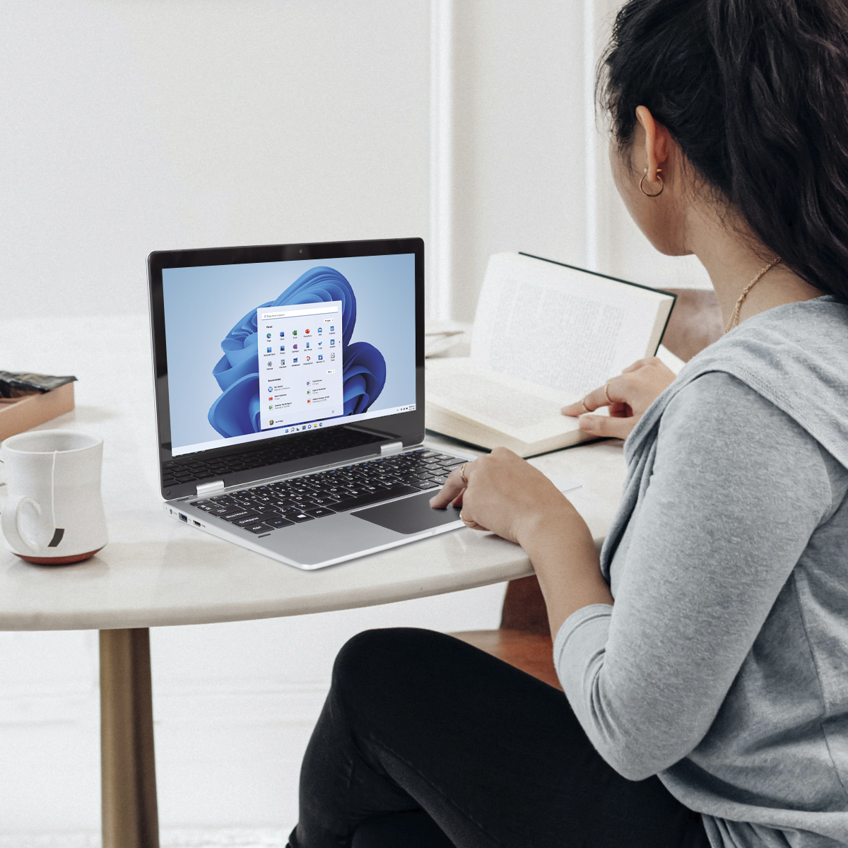 Eine Frau benutzt einen Laptop, liest ein Buch, mit einer Tasse auf einem Tisch. Weißer Hintergrund.