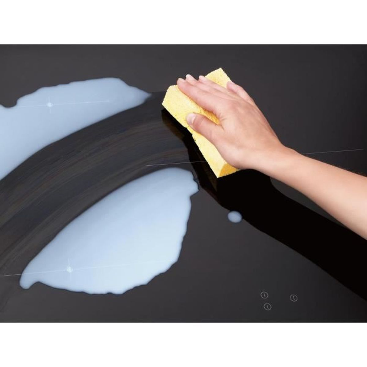 Ręka używająca żółtej gąbki do czyszczenia czarnej płyty kuchennej z rozlanym mlekiem.