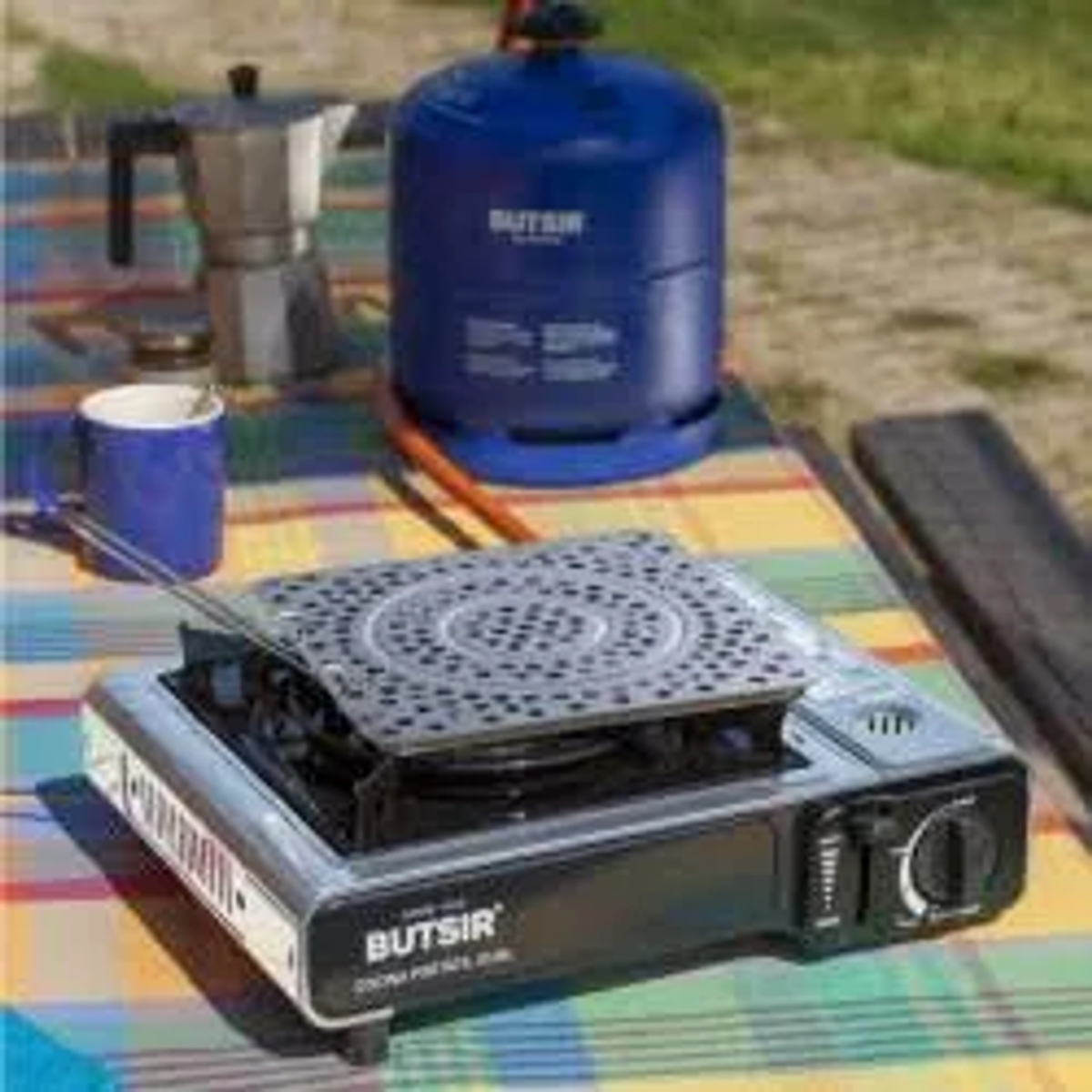 Una estufa de gas portátil con una botella de gas azul, una cafetera y una taza en una mesa.