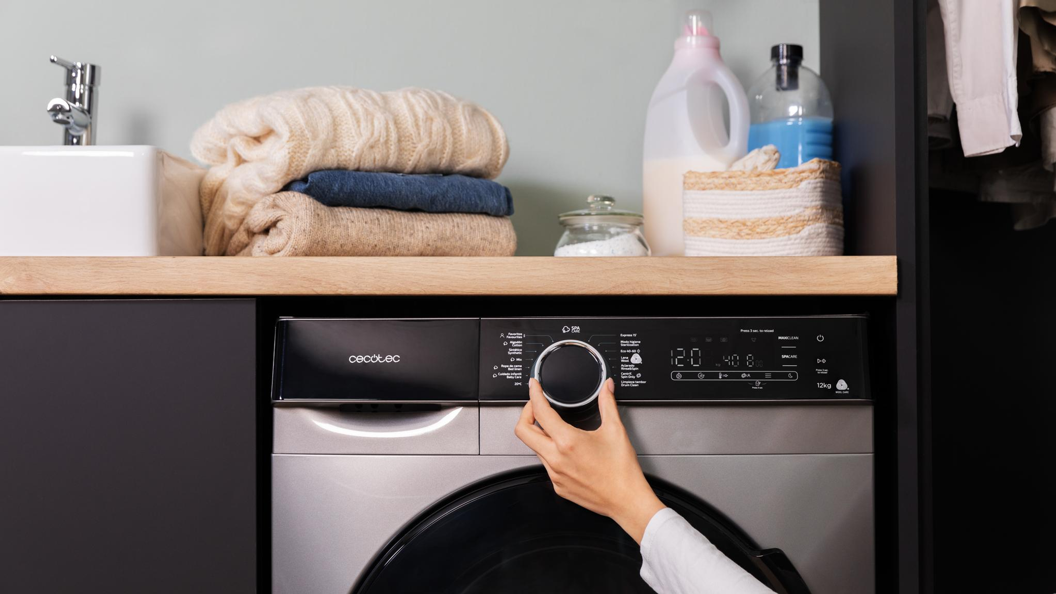 Dłoń osoby kręci pokrętłem pralki, w pobliżu inne przedmioty.