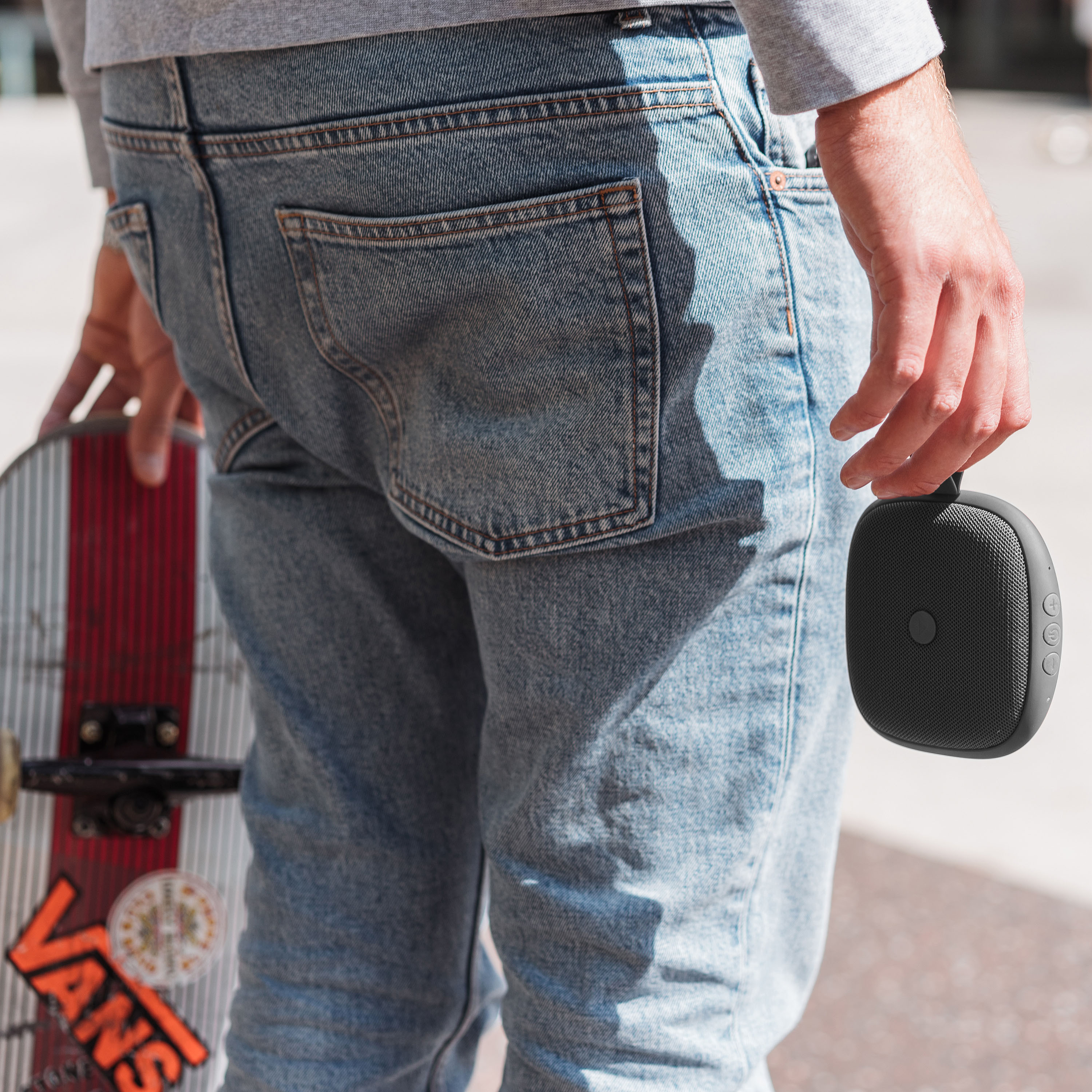 Mężczyzna trzymający czarny głośnik i deskorolkę. Jeansy i szara koszula.