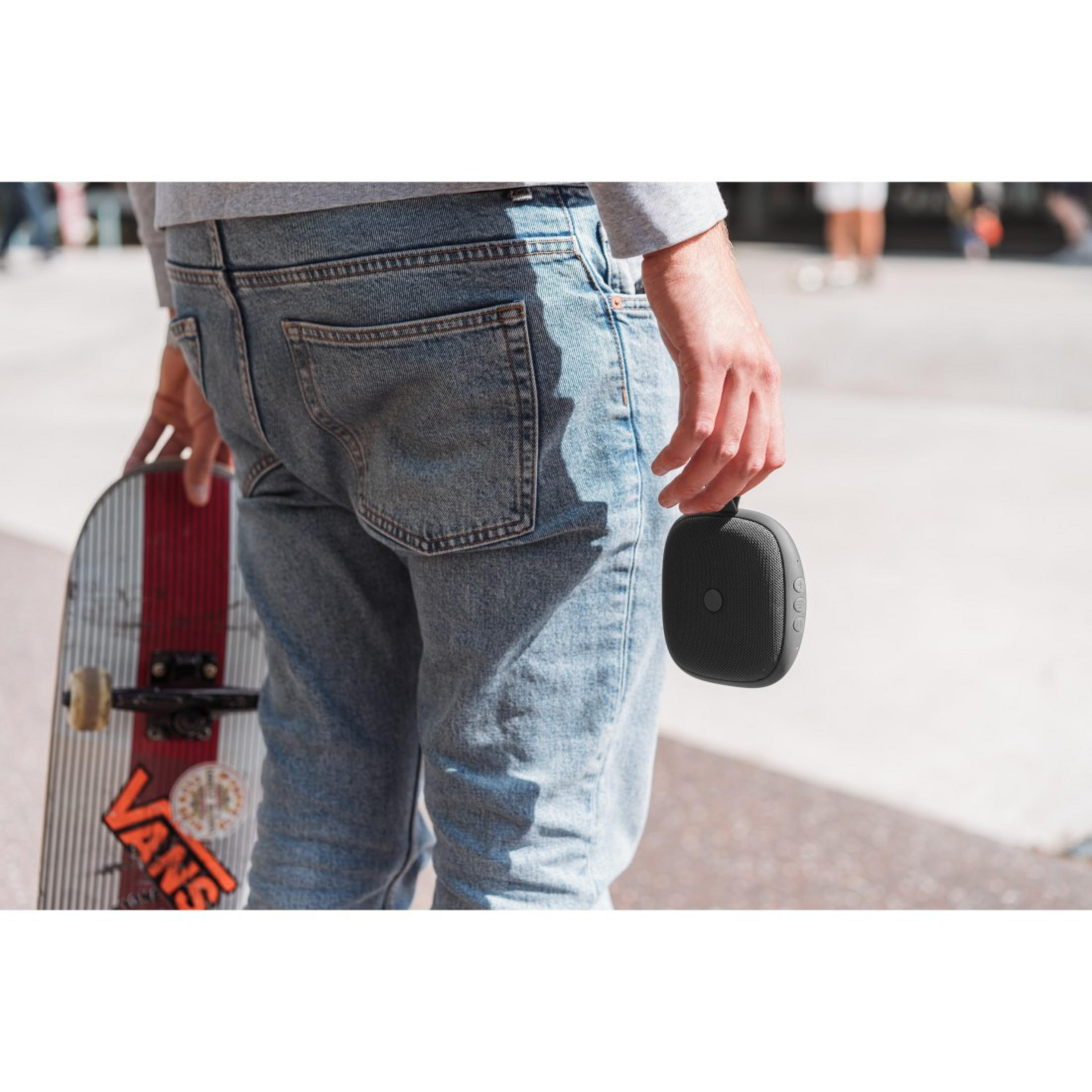 Osoba trzymająca czarny głośnik i deskorolkę z naklejkami. Miejsce akcji to plener.