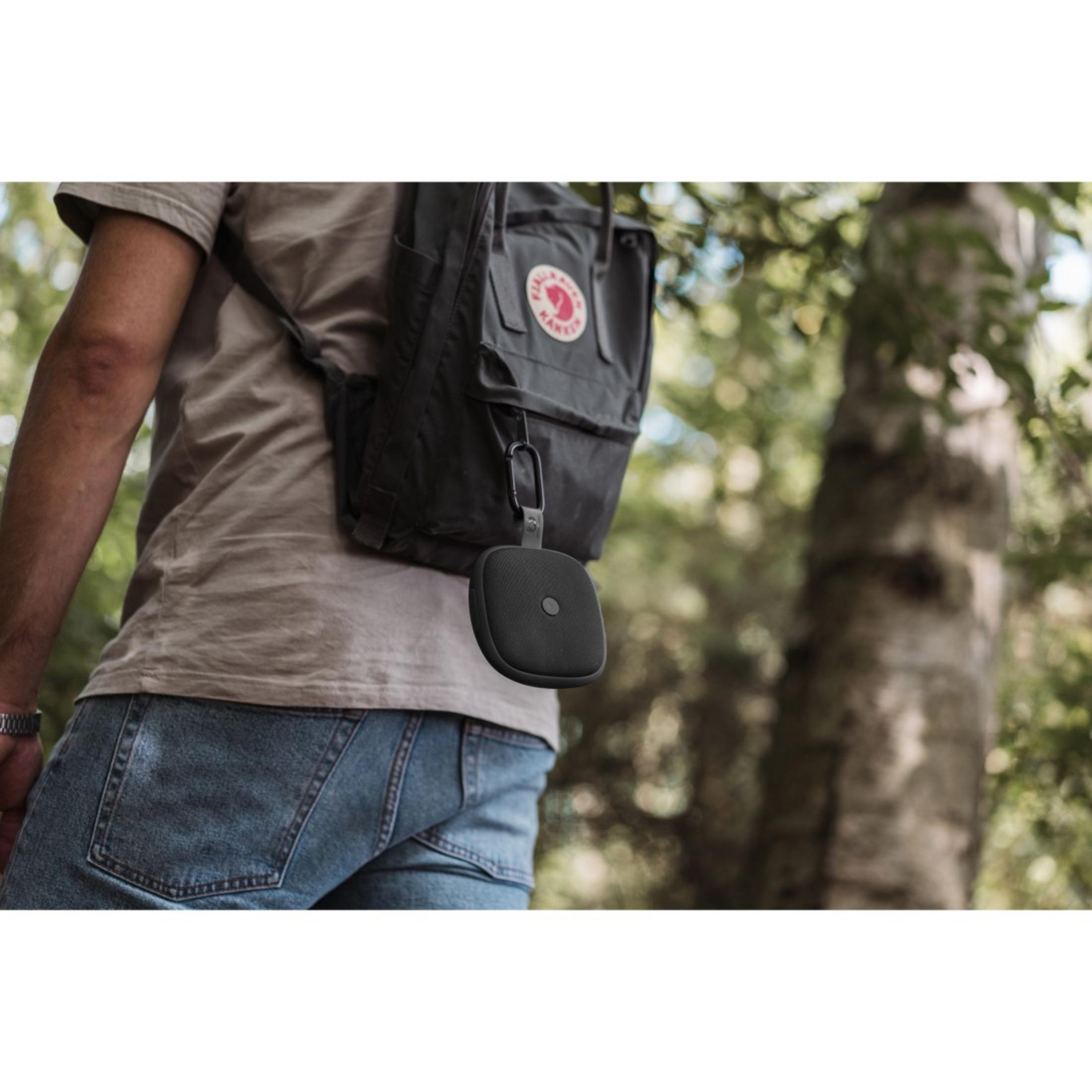 Osoba w zielonym otoczeniu z czarnym okrągłym głośnikiem przymocowanym do plecaka.