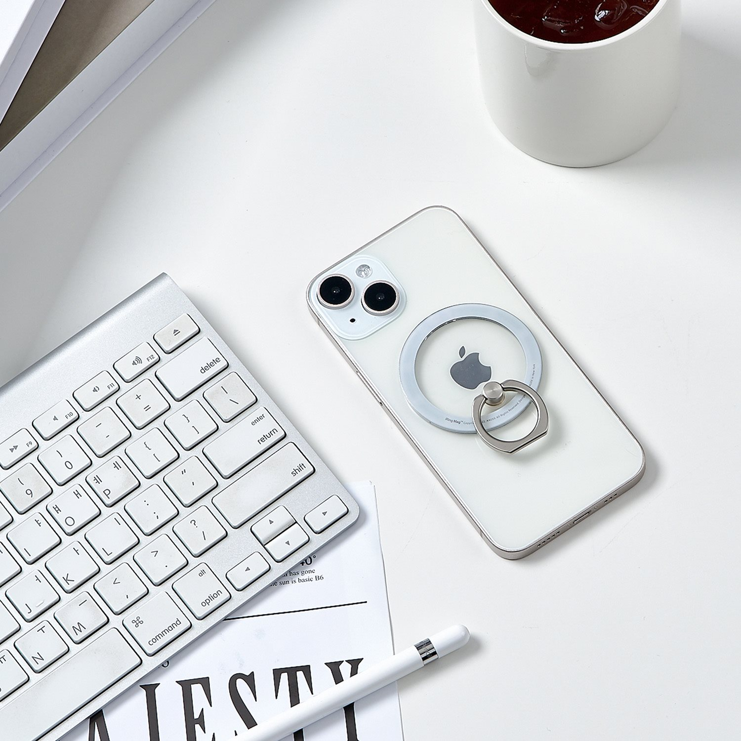 Ein weißes iPhone mit einem Ringhalter auf einem weißen Schreibtisch, zusammen mit einer Tastatur, einem Stift und einer Kaffeetasse.