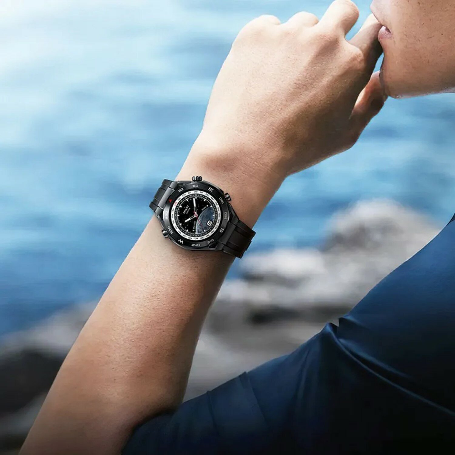 Ein Mann trägt eine schwarze Uhr am linken Handgelenk, blauer Wasserhintergrund.