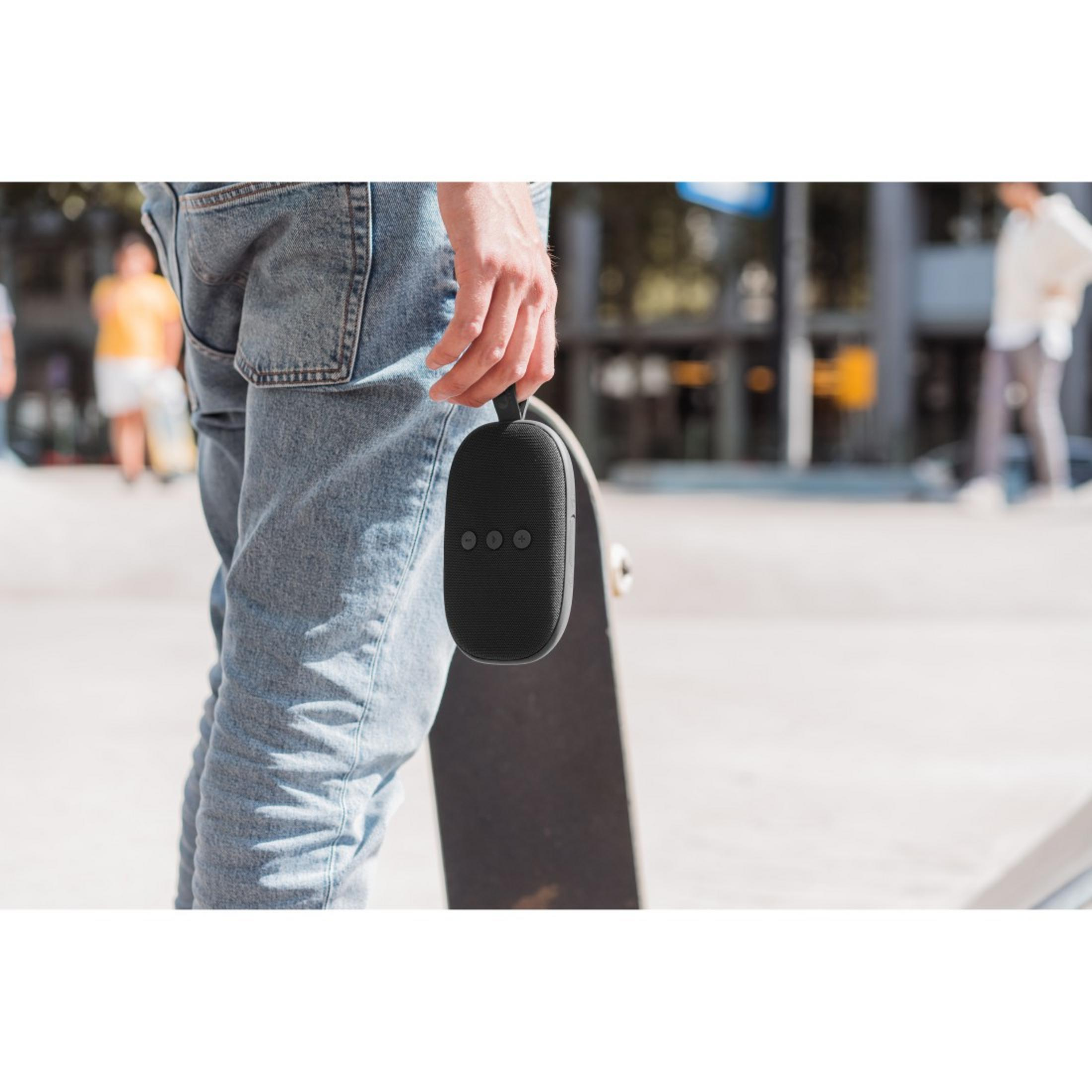 Een man die een zwarte speaker vasthoudt. Hij draagt een spijkerbroek en staat in een skatepark.