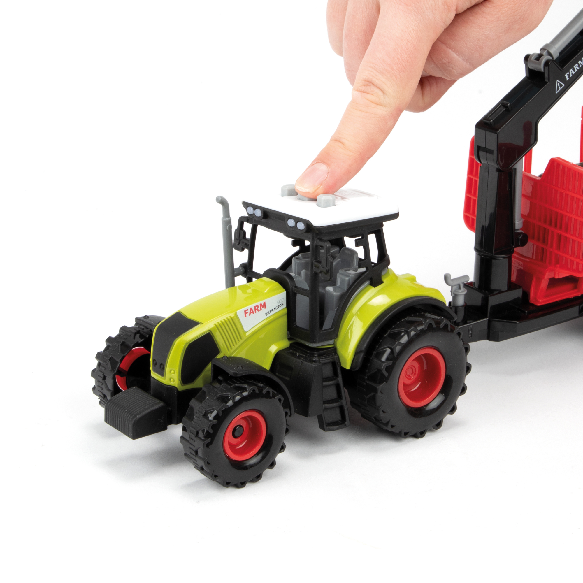 Ein Spielzeugtraktor mit dem Finger einer Person auf einem Knopf. Der Traktor ist grün und gelb, mit einem roten Anhänger.