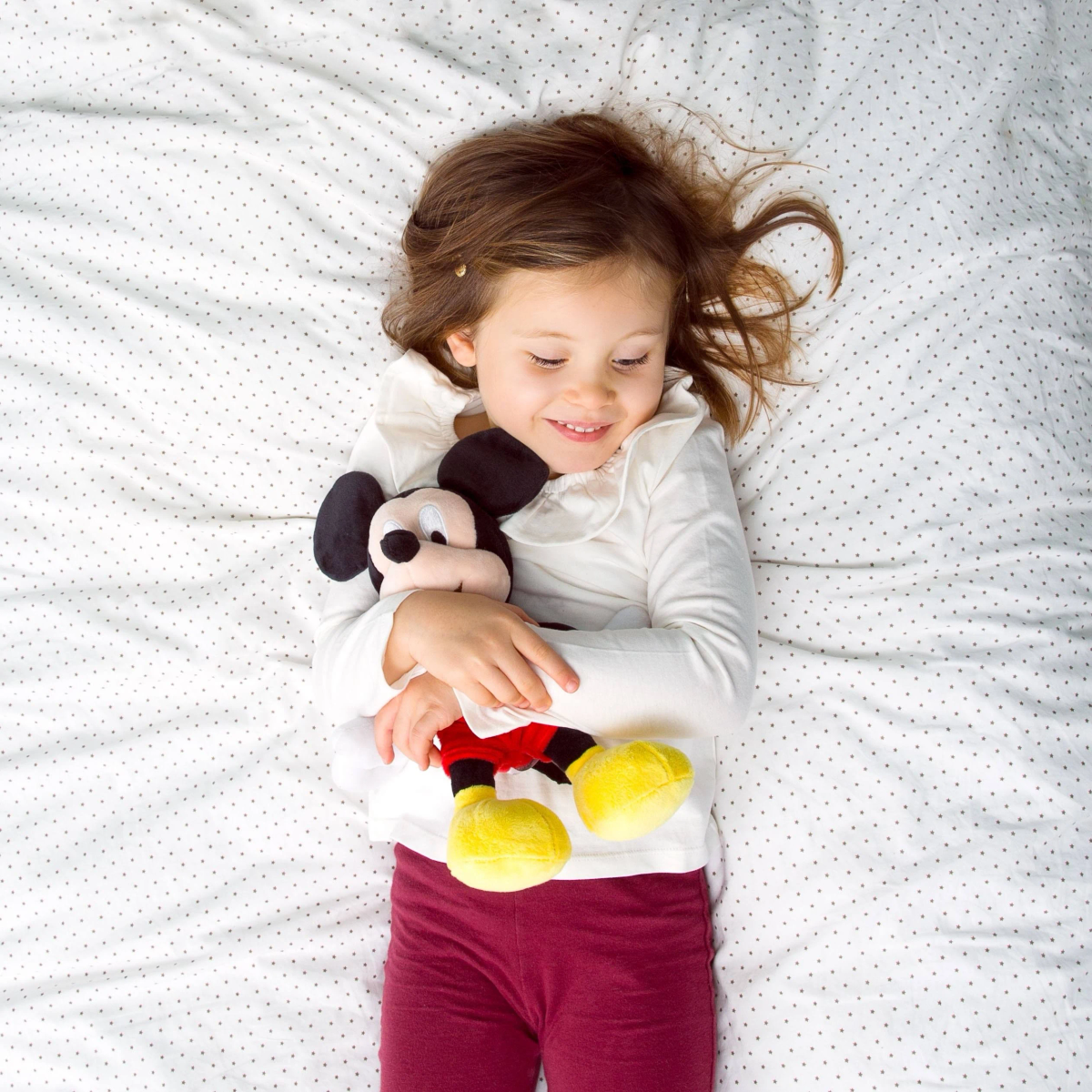 Ein Mädchen liegt auf dem Bett und umarmt ein Mickey-Mouse-Plüschtier. Sie lächelt.