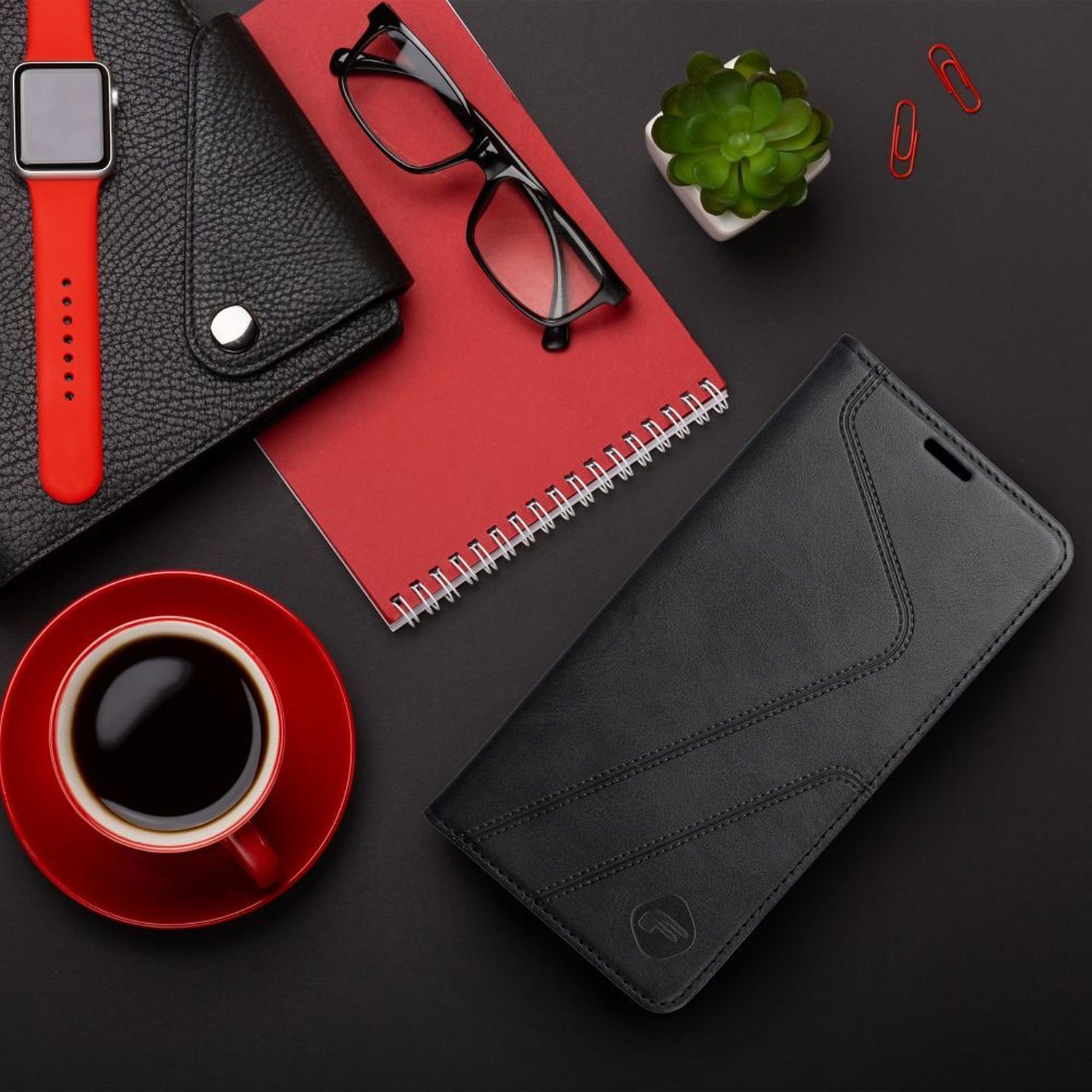 Una tazza di caffè rossa, un portafoglio nero, occhiali, un blocco note, uno smartwatch e una custodia per telefono su nero.