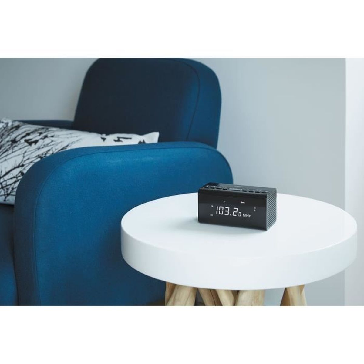 Czarny radioodtwarzacz cyfrowy na białym stole obok niebieskiej sofy. Widoczna poduszka dekoracyjna.