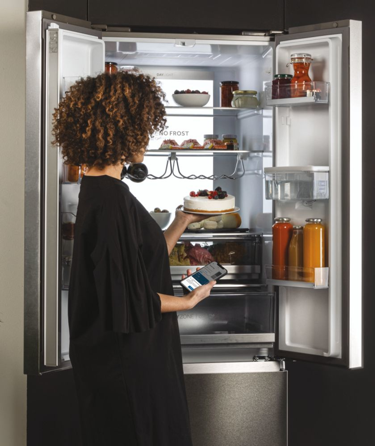 Mujer abriendo un refrigerador, sosteniendo un pastel y un teléfono. El refrigerador está lleno de comida y bebidas.