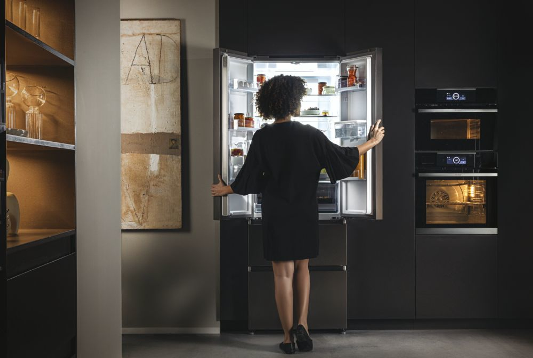 Mujer en vestido negro abriendo refrigerador en cocina moderna. Gabinetes oscuros, hornos.