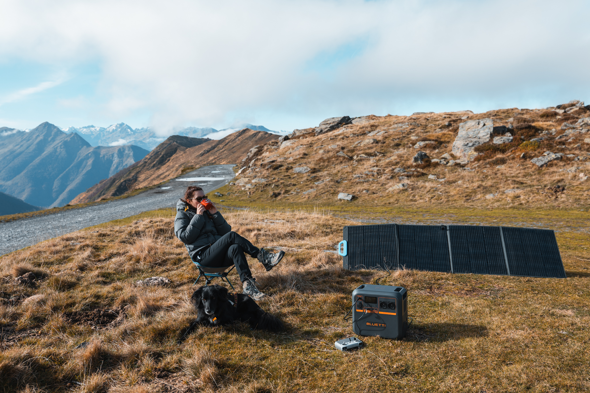 Osoba siedząca, pijąca, z psem na wzgórzu. Panel słoneczny i elektrownia w pobliżu. Góry w oddali.