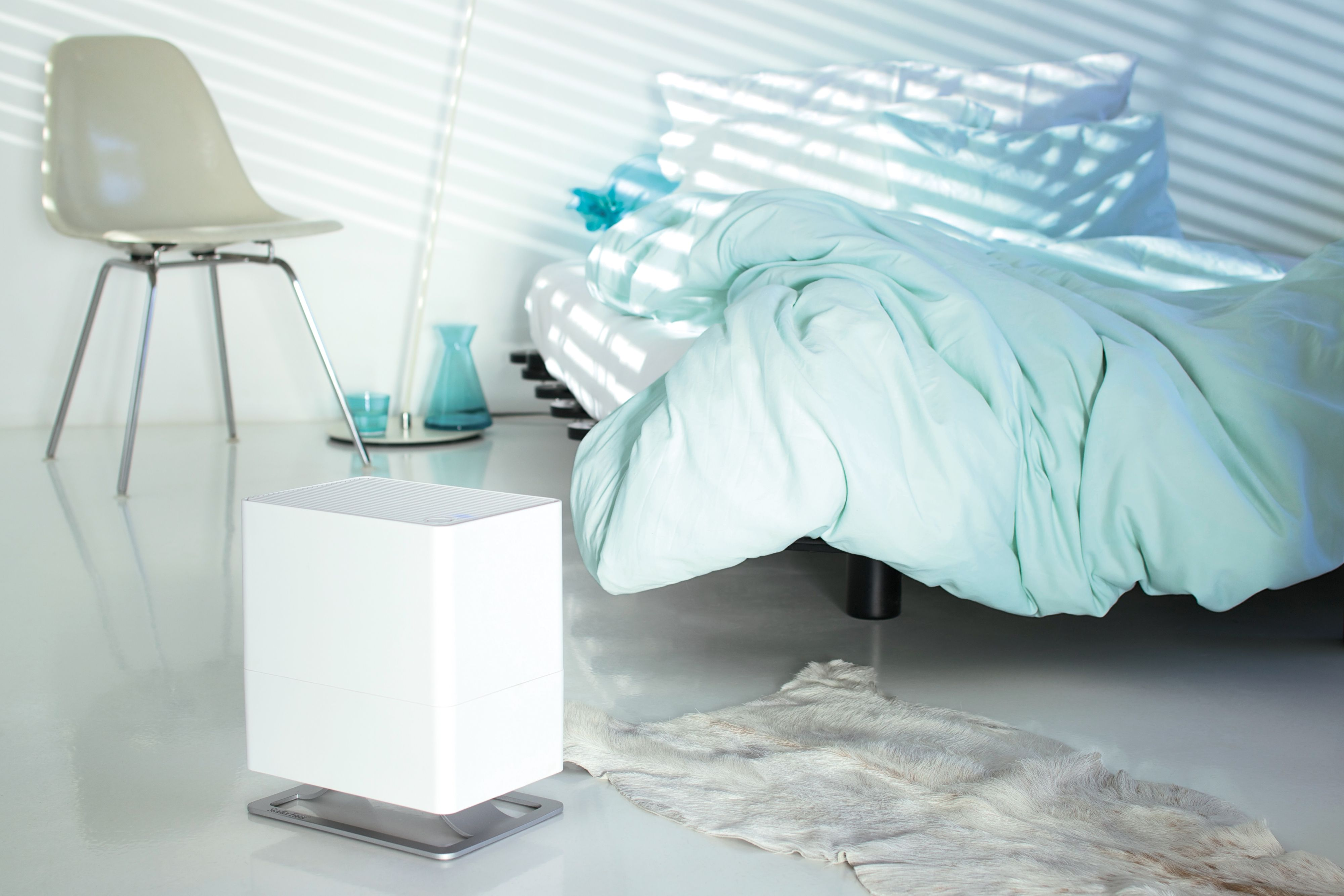 A white humidifier on a stand by a bed with a light blue blanket. A chair is on the left.