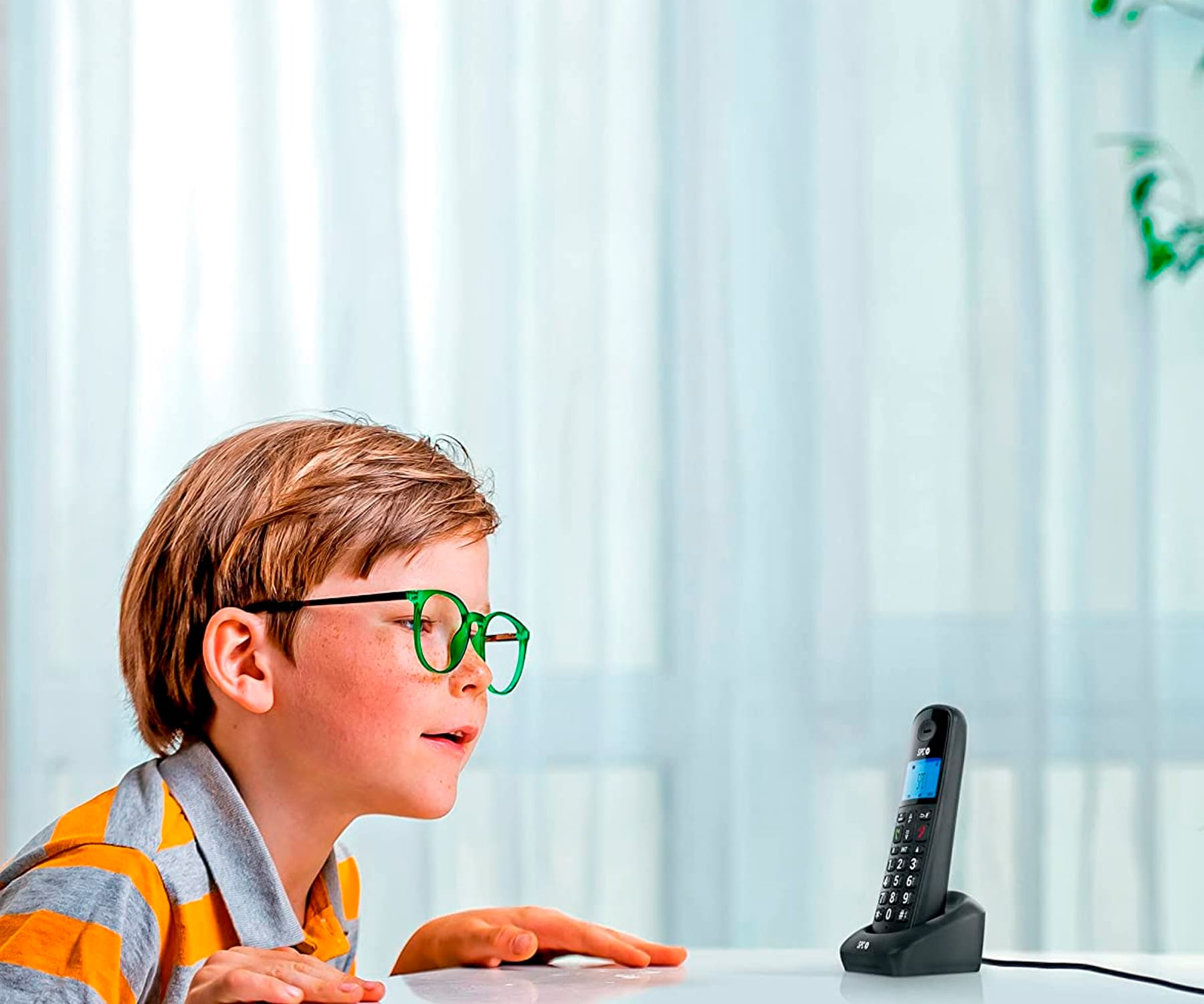 Ragazzo che guarda un telefono su un tavolo. Telefono in una base. Tende bianche sullo sfondo.
