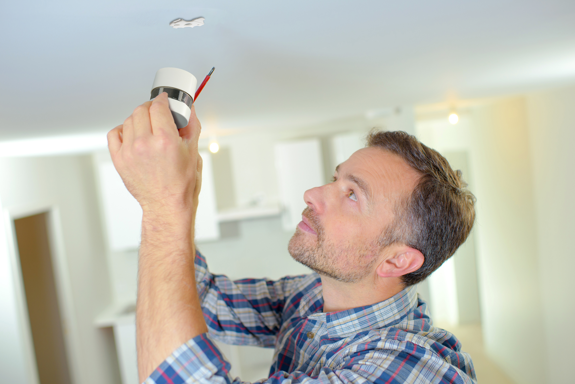 Mężczyzna instaluje czujnik dymu na białym suficie. Pokój w tle.