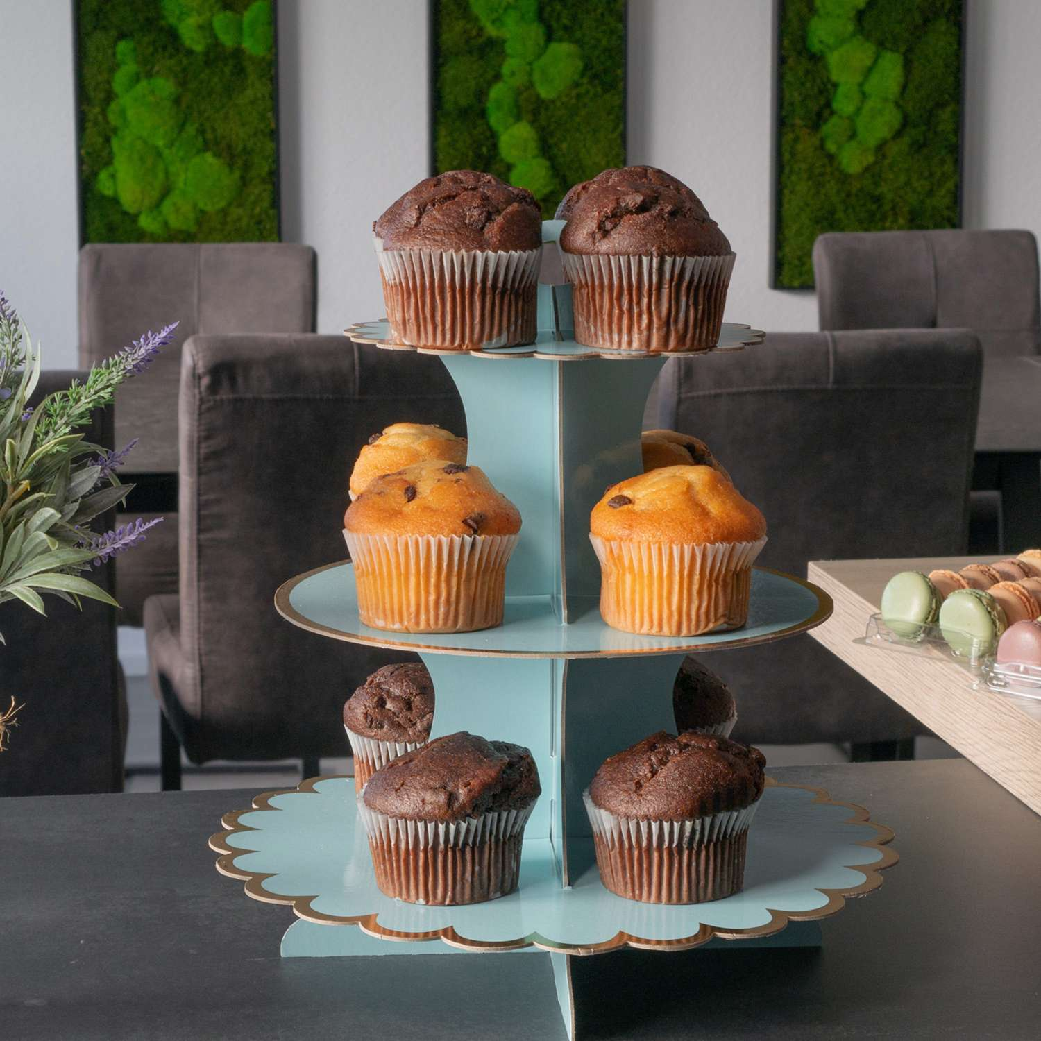 Ein dreistöckiger Cupcake-Ständer mit Schoko- und Vanille-Cupcakes. Hintergrund mit Stühlen.