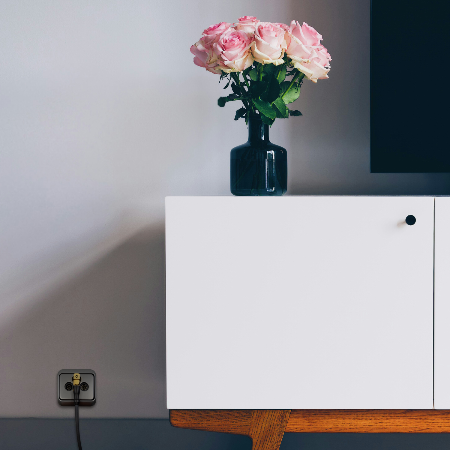 Eine Vase mit rosa Rosen auf einem weißen Schrank mit einem schwarzen Fernseher und einer Steckdose.