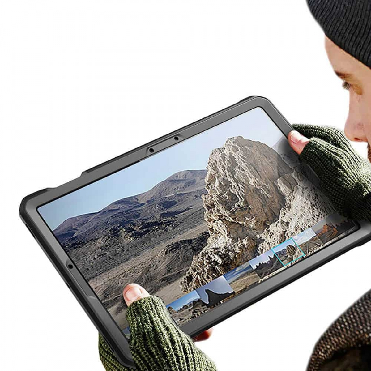 Eine Person hält ein Tablet, das eine Berglandschaft zeigt. Sie trägt Handschuhe und eine Mütze.