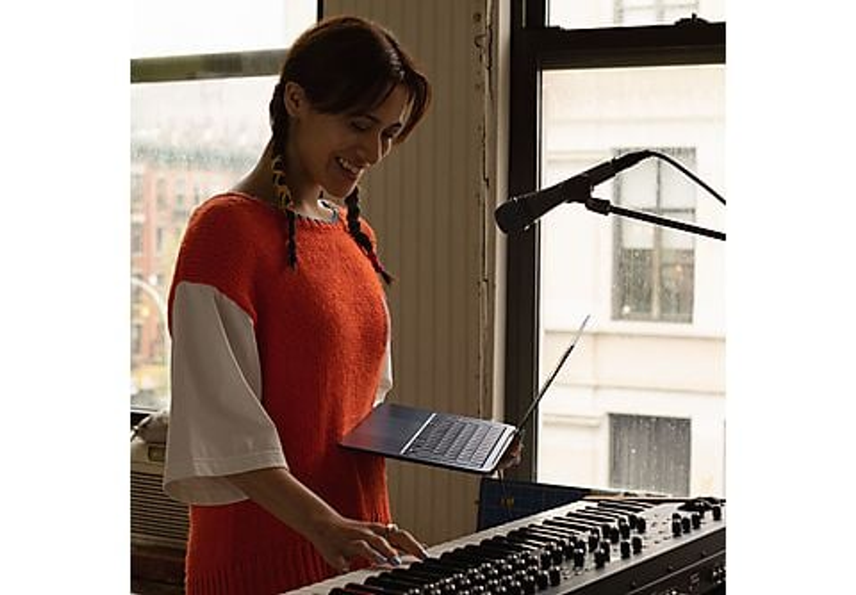 Una donna suona la tastiera, sorridendo, in una stanza con una finestra e un'asta per microfono.