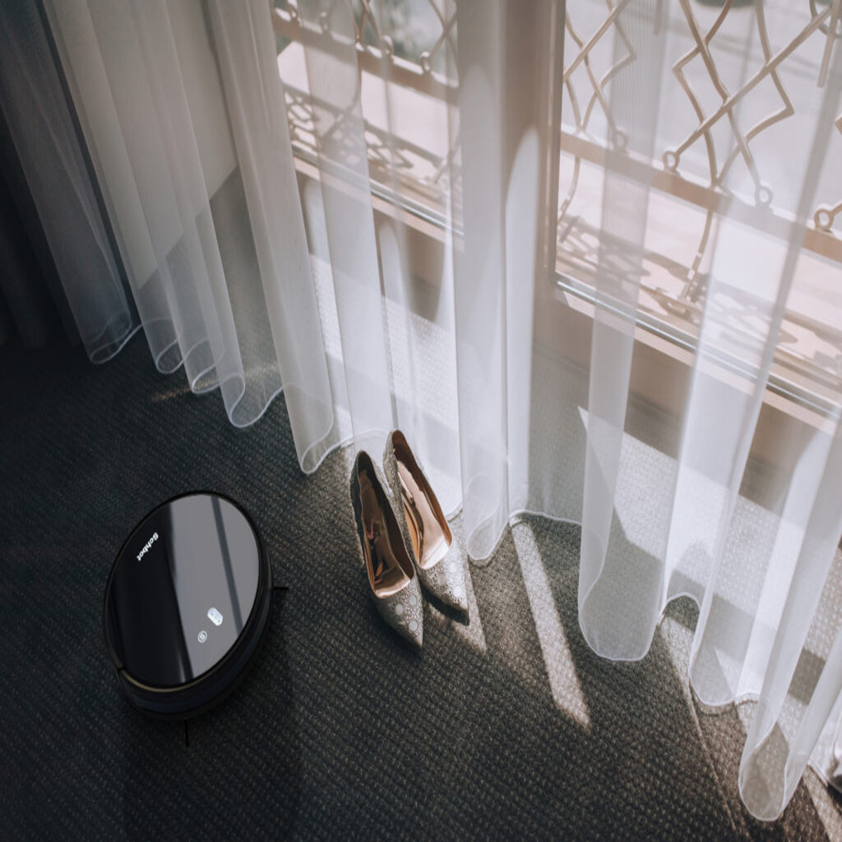 Ein Roboter-Staubsauger, in der Nähe von Schuhen und Vorhängen. Sonnenlicht durch das Fenster.