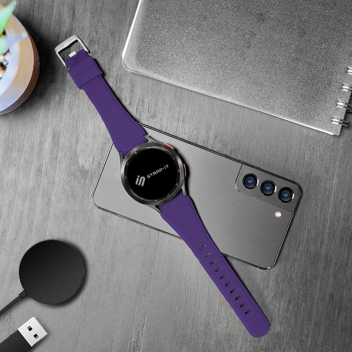 Smartwatch mit lila Armband auf einem kabellosen Ladegerät. Handy und Notizbuch daneben.