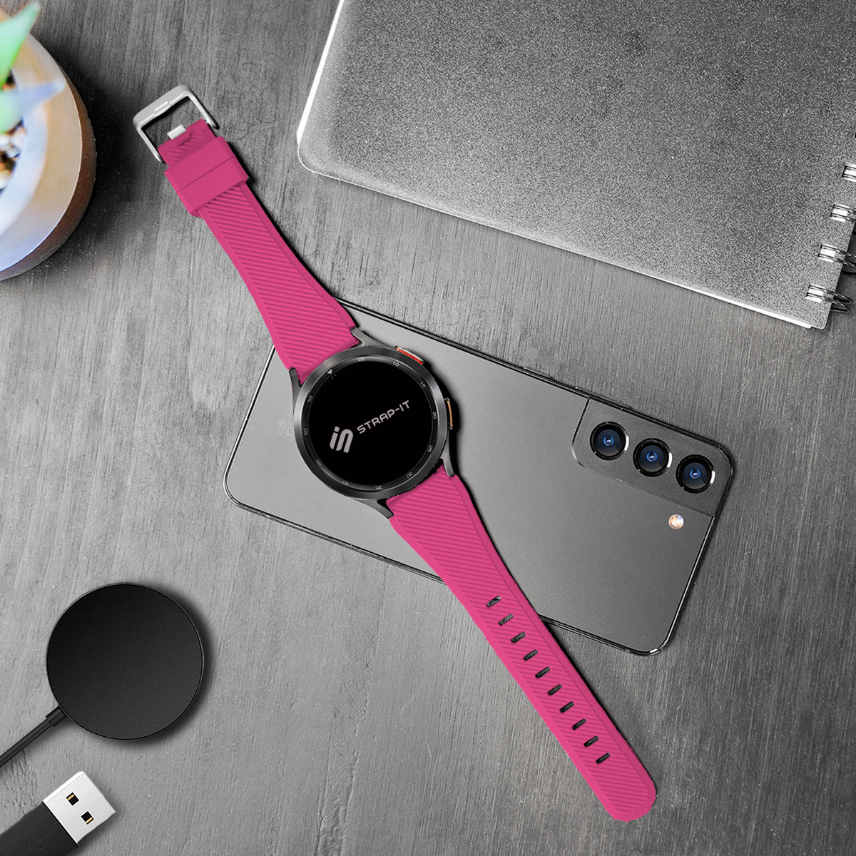 Smartwatch mit pinkfarbenem Armband, ein Telefon und ein Ladepad auf Holzoberfläche. Text: Strap-IT.