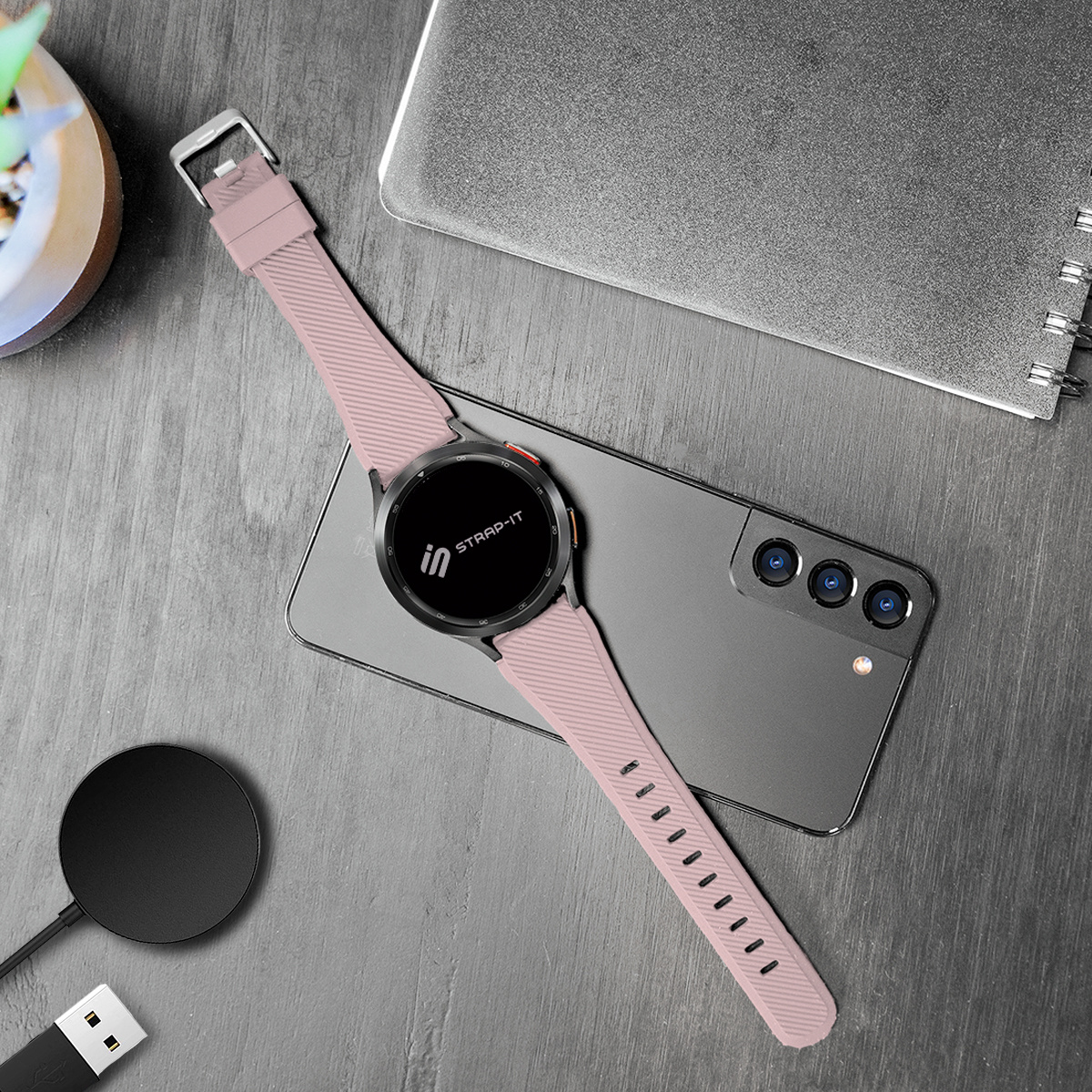 Eine Smartwatch mit rosa Armband liegt auf einem grauen Telefon. Es gibt eine Ladestation und einen Holztisch.