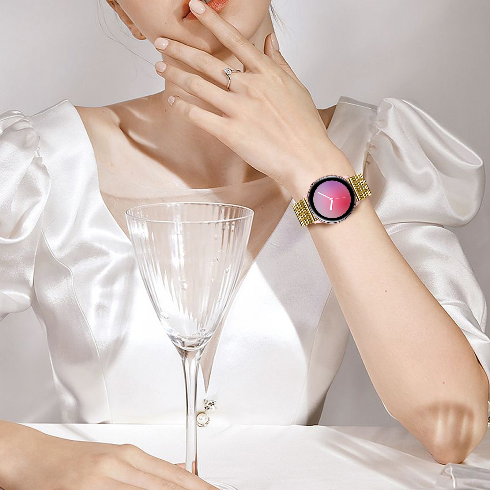 Frau mit goldener Uhr. Sie trägt ein weißes Kleid und sitzt am Tisch.