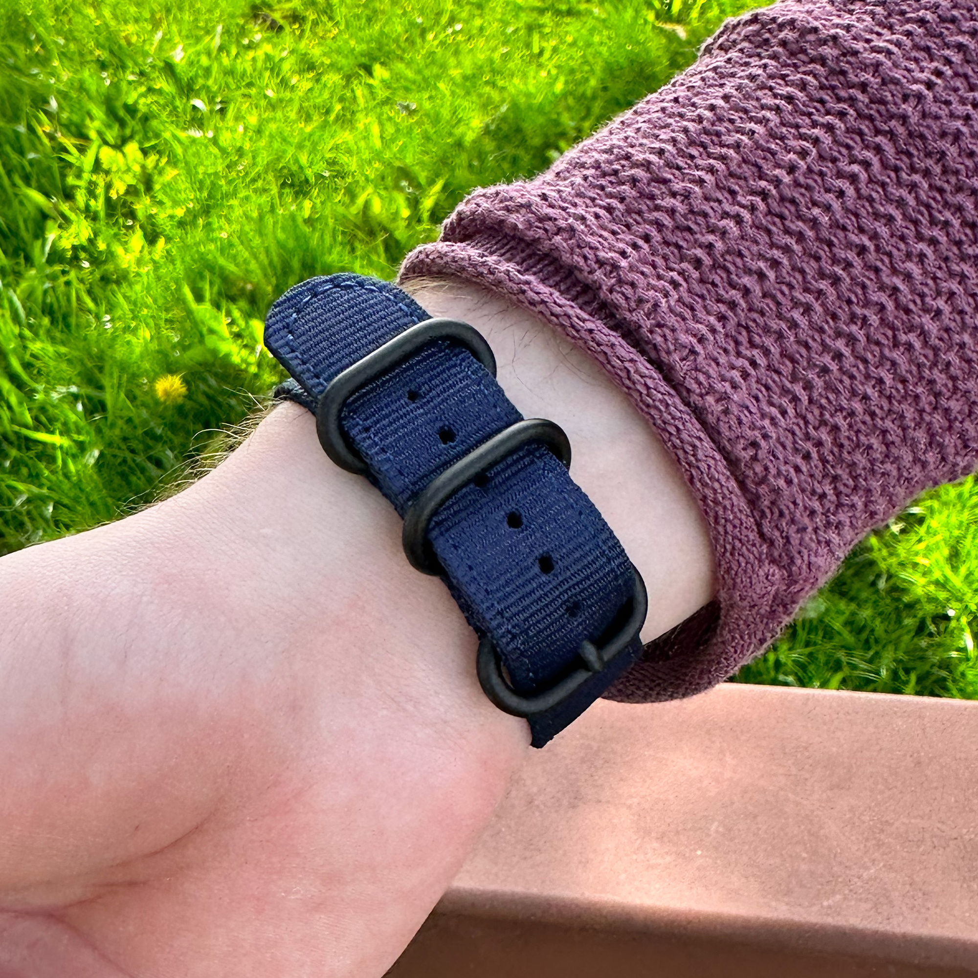 Ein Handgelenk mit einer Uhr. Die Uhr hat ein dunkelblaues Armband und schwarze Hardware. Der Hintergrund ist grünes Gras.