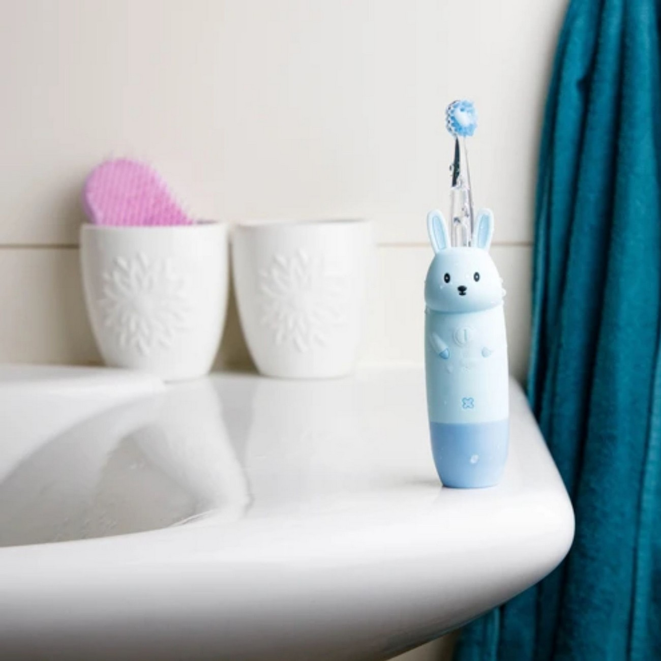 Cepillo de dientes azul y blanco con cabeza de conejito. Lavabo blanco y dos tazas al fondo.