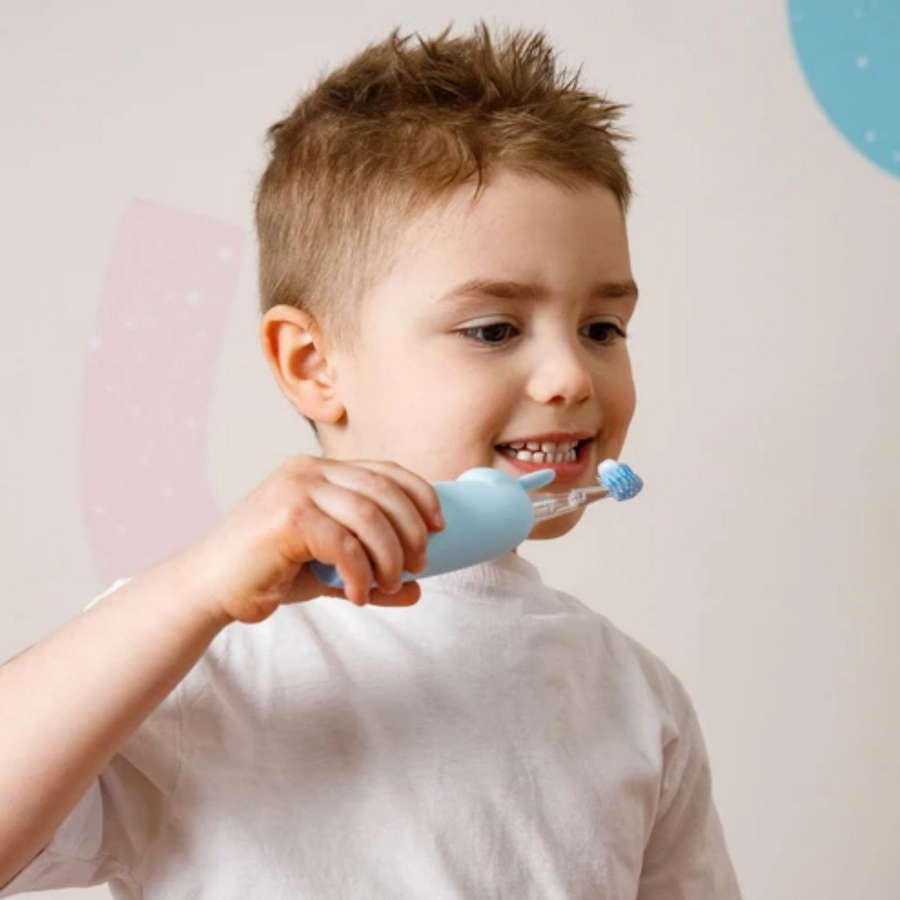 Un niño se cepilla los dientes con un cepillo eléctrico azul, sonriendo, con una camisa blanca.