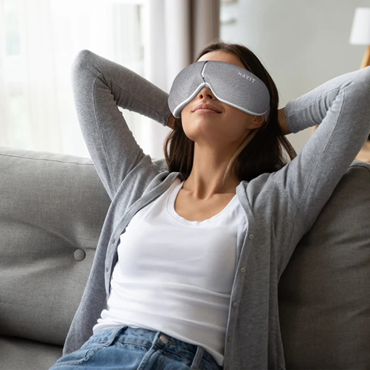 Mujer con antifaz gris en sofá gris, manos en la cabeza, pose relajada.