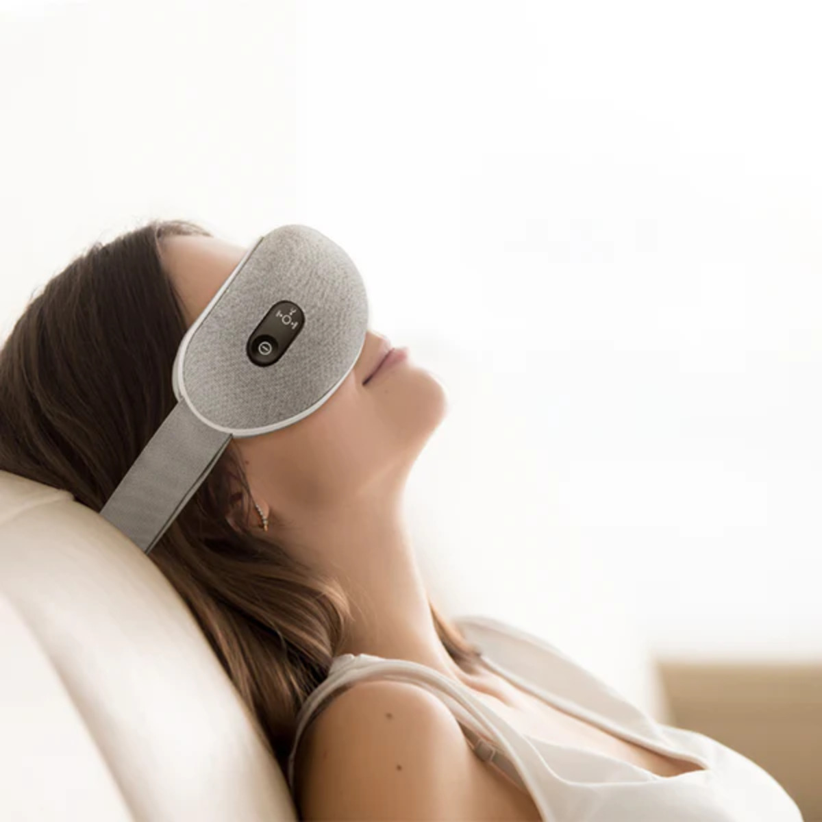 Una mujer con una máscara para los ojos gris, acostada. Cierra los ojos relajada, fondo blanco.