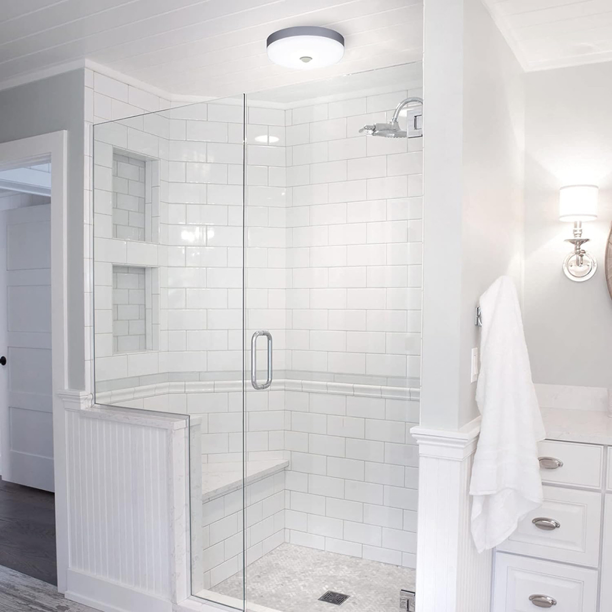 Badezimmerdusche mit weißen Fliesen, Glastür und einer Sitzbank. Gut beleuchtet.