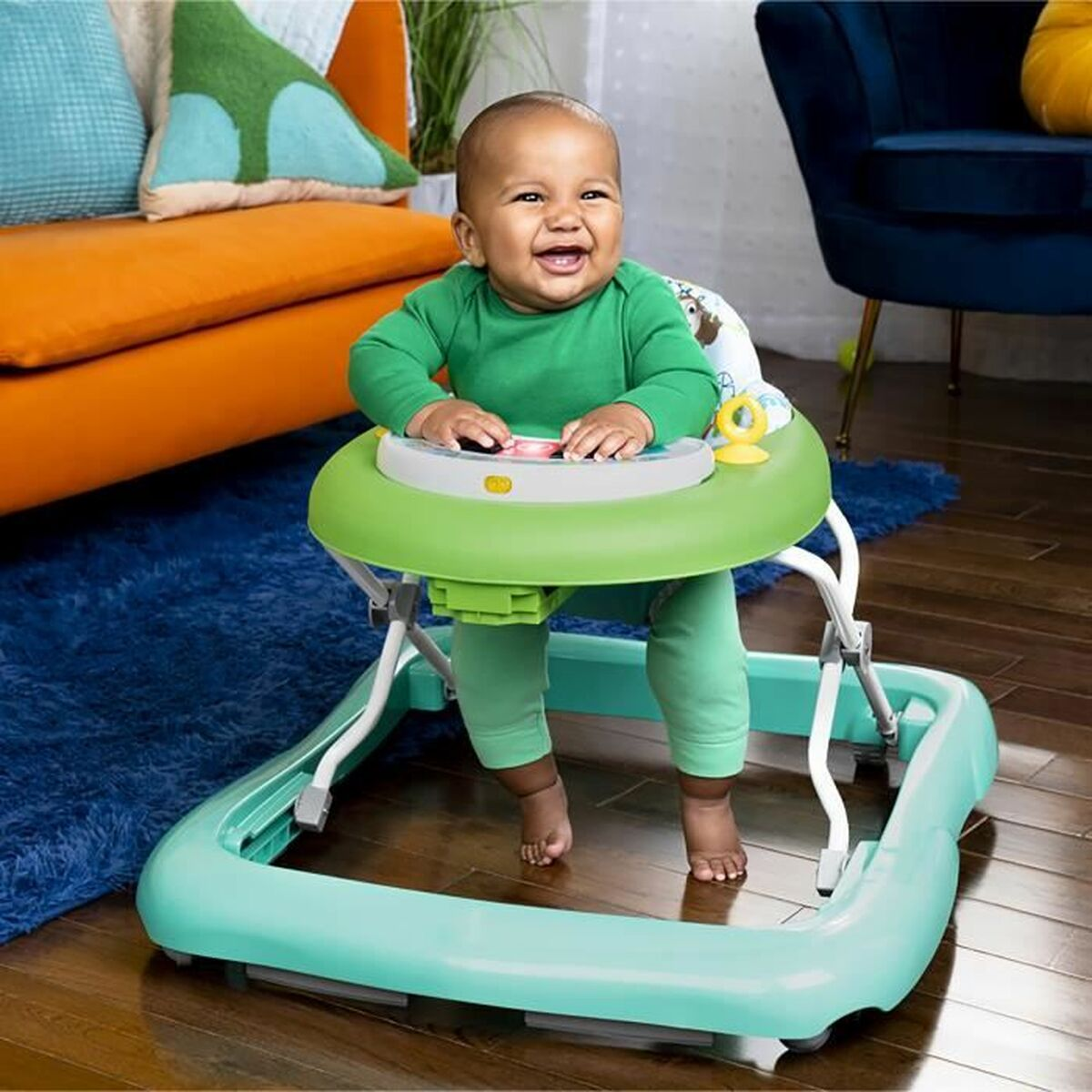 Ein Baby in grünem Outfit sitzt in einem Baby-Walker auf einem blauen Teppich.