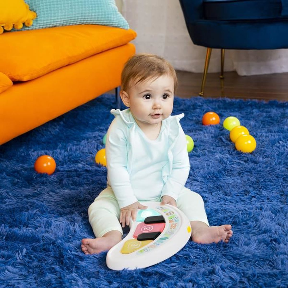 Baby sitzt auf blauem Teppich und spielt mit einem Klavierspielzeug. Orangefarbene Bälle sind sichtbar. Verschwommener Hintergrund.