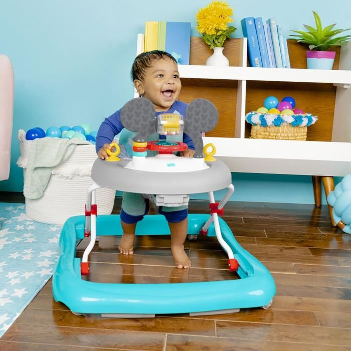 Un niño pequeño en un andador sonríe. El andador es turquesa y gris. El fondo incluye juguetes y muebles.