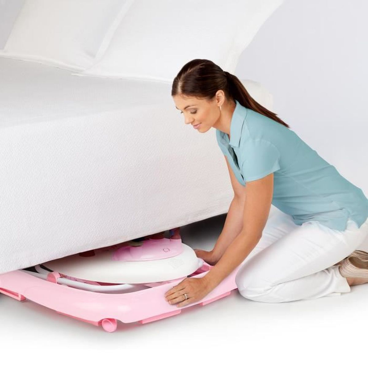 Eine Frau kniet und platziert eine rosa Babyspielmatte unter einem Bett. Das Bett hat weiße Laken.