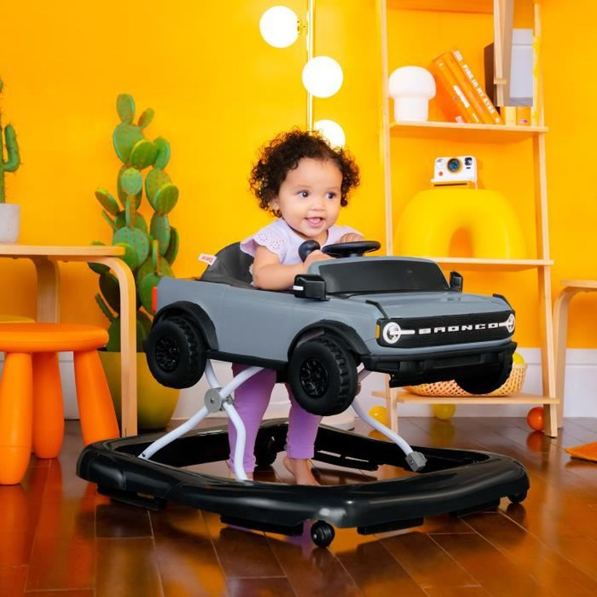 Baby in grauem Walker, gelber Hintergrund, kleiner orangener Tisch, Pflanzen im Regal.