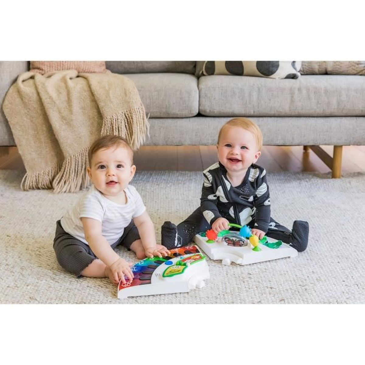 Zwei Babys sitzen auf einem Teppich und spielen mit Spielzeug. Eine Couch steht dahinter.