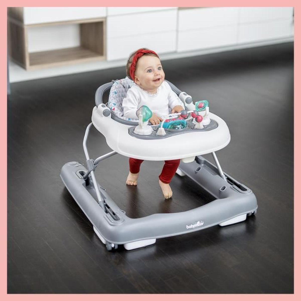 Ein Baby in einem Gehwagen lächelt, mit rosa Hintergrund und einem Spielbereich vorne.