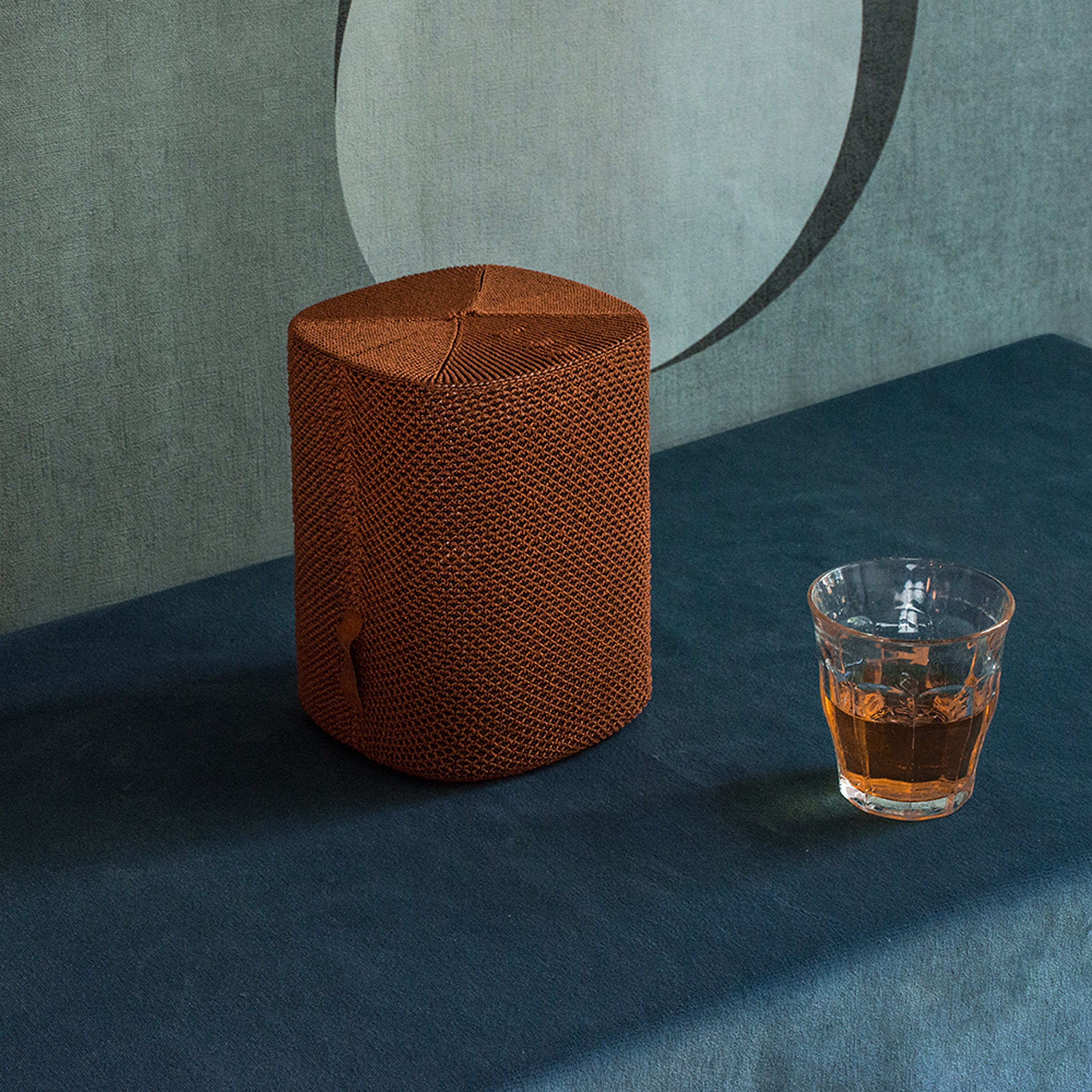 Un tabouret orange tissé et un verre de liquide ambré reposent sur une surface bleue.