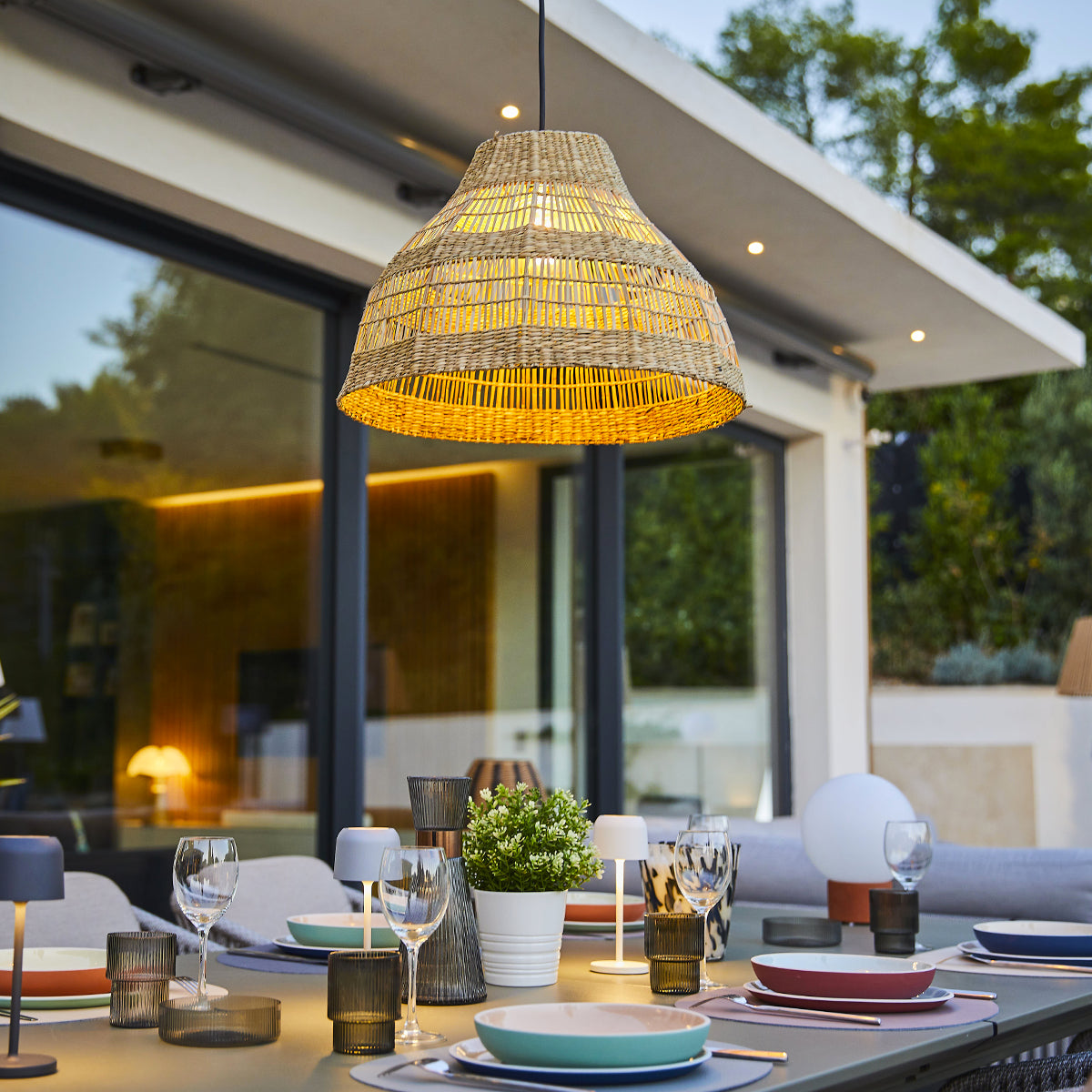 Essplatz im Freien mit gewebter Lampe, Tisch und Geschirr. Eine moderne, entspannte Atmosphäre.